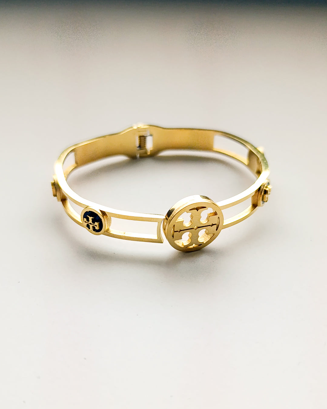 A gold-colored metal ankle cuff bracelet with a sleek and minimalist design, featuring a brand logo.